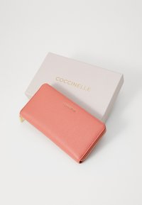 Koraal leren portemonnee met een gestructureerde afwerking, gouden hardware en een ritssluiting, geplaatst op een lichtroze doos met gouden letters.
