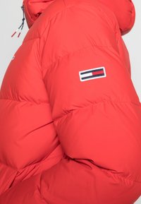 Rød puffjakke med en quiltet tekstur, featuring et mærke med blå, hvide og røde vandrette striber på ærmet. Hætte-design.
