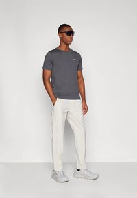 Grå t-shirt med korta ärmar och en liten logotyp, kombinerad med vita byxor och grå sneakers. Modellen bär mörka solglasögon.