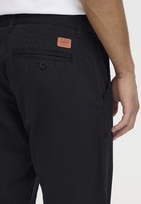 Pantalons en mélange de coton noir avec une poche latérale, une poche arrière fermée par un bouton et une étiquette en cuir texturé. Design ajusté, tissu léger.