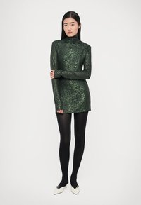 Zöld, bordázott mini ruha magas gallérral, hosszú ujjakkal és texturált, csillogó felülettel. Fekete harisnyával és fehér magassarkúval viselve.