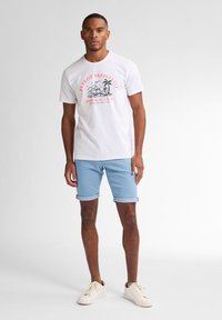 Camiseta blanca de algodón con un gráfico de color de olas y palmeras, combinada con shorts de mezclilla azul claro arremangados y zapatillas blancas.