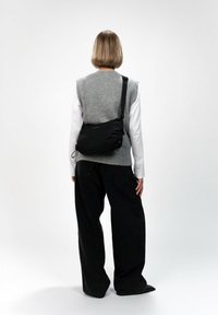 Borsa a tracolla nera con texture arricciata, tracolla regolabile e dettagli del logo sottili, indossata sopra un gilet grigio e jeans neri a gamba larga.