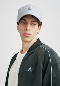 Boné cinza com um design estruturado e logotipo prateado, combinado com uma jaqueta preta com detalhes azul-claro e uma t-shirt branca.