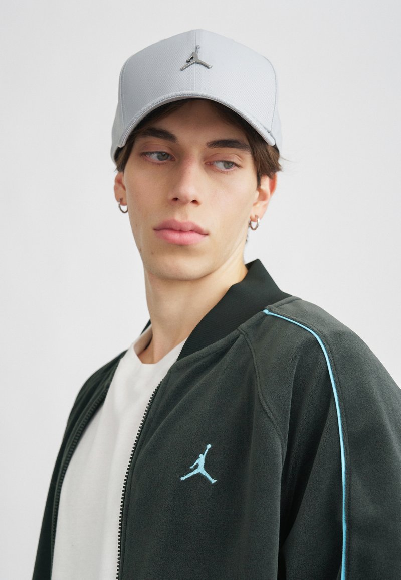 Boné cinza com um design estruturado e logotipo prateado, combinado com uma jaqueta preta com detalhes azul-claro e uma t-shirt branca.