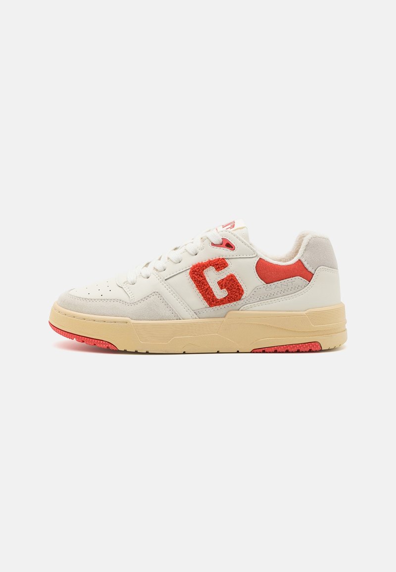 Baskets blanches et beiges avec des accents rouges, un grand "G" rouge texturé sur le côté, et une semelle beige avec des détails de la semelle rouges.