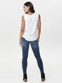 Top bianco senza maniche con pizzo sulle spalle, abbinato a jeans aderenti blu scuro. La modella ha i capelli lunghi e scuri, in piedi di profilo.