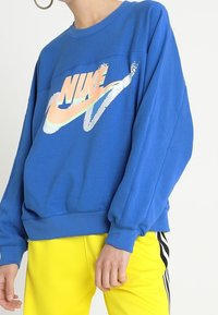 Blå oversized sweatshirt med rund hals och långa ärmar som har en flerfärgad Nike-logga i mitten. I kombination med gula byxor.