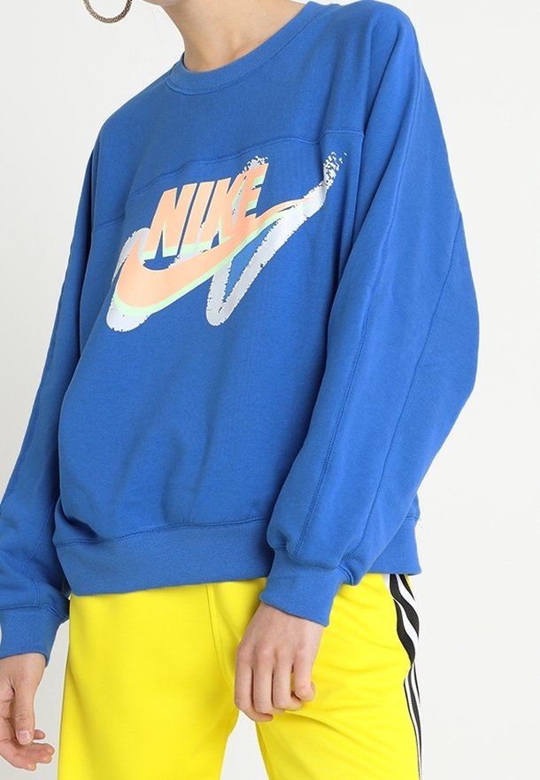 Blå oversized sweatshirt med rund hals och långa ärmar som har en flerfärgad Nike-logga i mitten. I kombination med gula byxor.