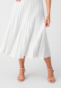 Jupe midi plissée blanche en tissu doux, dotée de plis verticaux. Portée avec des sandales à talons aiguilles nude à lanières, mettant en valeur une texture lisse.