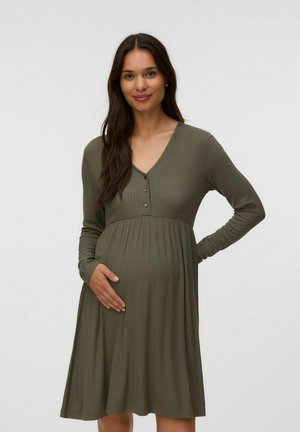 Zwangere vrouw met lang bruin haar draagt een geribde, olijfgroene jurk met lange mouwen, glimlacht en houdt met één hand haar buik vast. Effen achtergrond.