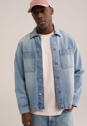 Man draagt lichtblauwe spijkerjack over een lichtroze shirt en bijpassende roze pet, staande tegen een effen beige achtergrond.