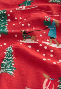 Tessuto rosso con un motivo natalizio che presenta alberi verdi, neve, persone e un cane, tutti illustrati in vari colori e scene giocose.