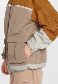 Brun och beige färgblockerad jacka med dragkedjefickor, elastiska ärmmuddar och en grå fåll. Tillverkad av lätt, vattentåligt material.