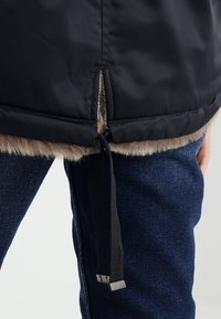 Bretelle à cordon suspendue à l'ourlet d'une veste noire doublée de fourrure, portée sur un jean en denim bleu foncé.