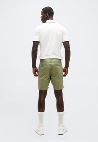 Ανδρας που στέκεται γυρισμένος προς τα πίσω, φοράει άσπρη κοντομάνικη πόλο, πράσινες βερμούδες, λευκές κάλτσες και λευκά αθλητικά παπούτσια σε απλό φόντο.