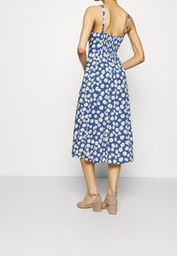 Robe d'été bleue avec des motifs de marguerites blanches, corsage froncé et jupe jusqu'aux genoux. Associée à des sandales à talons bloc beiges.