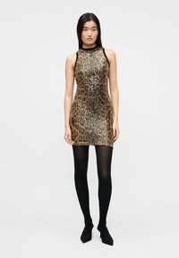 Γυναίκα που φοράει μίνι φόρεμα χωρίς μανίκια με παγιέτες σε χρυσό και μαύρο λεοπάρ μοτίβο, με μαύρο καλσόν και μαύρες γόβες, στέκεται μπροστά σε απλό φόντο.