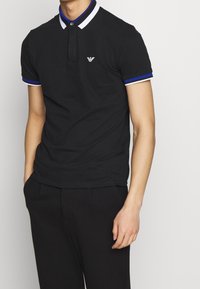 Man i svart kortärmad polotröja med vita och blå ränder på kragen och ärmarna, står med en hand i fickan.