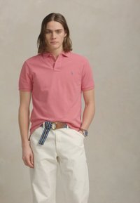 Jauns vīrietis ar plecu garuma matiem, ģērbts rozā polo kreklā, baltās biksēs, ar svītrainu jostu un rokas pulksteni, stāv ar vienu roku kabatā.