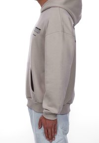 Sweat à capuche gris clair avec une poche kangourou, des poignets côtelés et des détails de logo en relief sur la poitrine et la manche. Texture lisse.