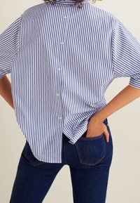 Chemise à manches courtes rayée bleu et blanc avec un col montant, une patte de boutonnage sur le devant, et une coupe légèrement ample, associée à un jean bleu foncé.