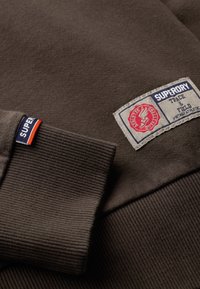 Sudadera marrón con puños acanalados, que presenta una etiqueta de tela con acentos rojos y azules. El material parece suave y texturizado.