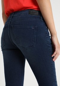 Jeans de mezclilla azul oscuro con un corte ajustado, que cuentan con bolsillos traseros y sutiles detalles de costura. El logo de la marca es visible en la cinturilla.