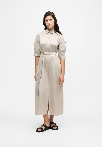 Femme portant une robe midi beige boutonnée avec une ceinture à la taille et des manches aux coudes, associée à des sandales noires à bouts ouverts, debout sur fond blanc.