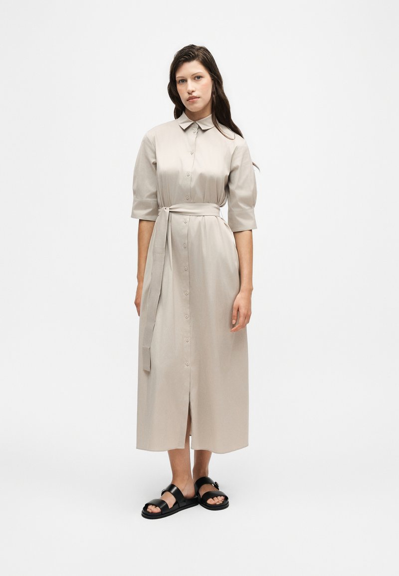 Femme portant une robe midi beige boutonnée avec une ceinture à la taille et des manches aux coudes, associée à des sandales noires à bouts ouverts, debout sur fond blanc.