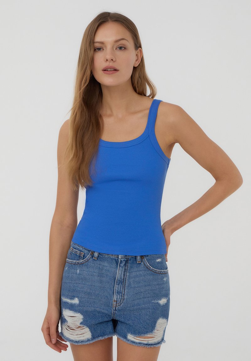 Canottiera blu a coste con spalline larghe, abbinata a pantaloni shorts in denim strappati. La canottiera ha una silhouette aderente e una texture morbida.