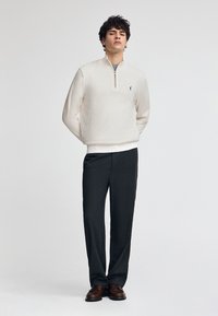 Ljusgrå, långärmad tröja med dragkedja i kragen, i kombination med mörkgrå byxor och bruna finskor. Enkelt logo på tröjan.