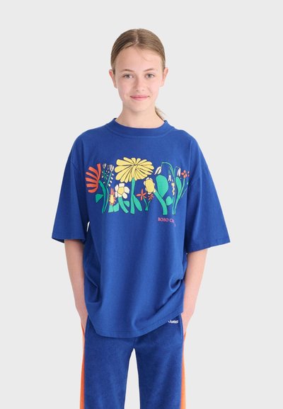 Junge Person trägt ein blaues, übergroßes T-Shirt mit buntem Blumenmuster und passende blaue Hose mit orangen Streifen, steht vor einem einfarbigen Hintergrund.