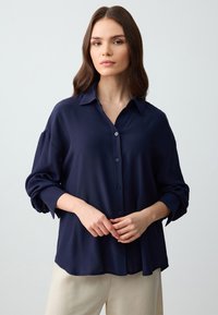 Marineblauwe blouse met knopen, gemaakt van zachte stof. Beschikt over een kraag, lange mouwen met opgerolde manchetten en een comfortabele pasvorm.