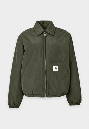 Personne portant une veste zippée vert foncé, un pantalon sombre, des chaussures à enfiler à carreaux et des lunettes de soleil, debout devant un fond uni.
