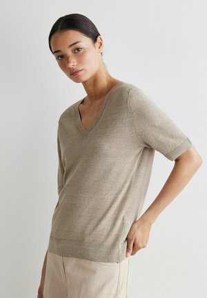 Kvinde med mørkt hår iført en beige v-neck kortærmet sweater og lyse bukser, står mod en hvid væg med hånden på hoften.