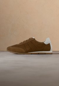 Baskets en daim marron avec languette de talon blanche et semelle en caoutchouc, présentées sur une surface et un fond neutres.