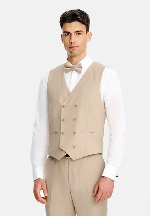 Młody mężczyzna ubrany w beżową kamizelkę dwurzędową, pasującą muszkę i spodnie, na białej koszuli, stojący na jednolitym tle.