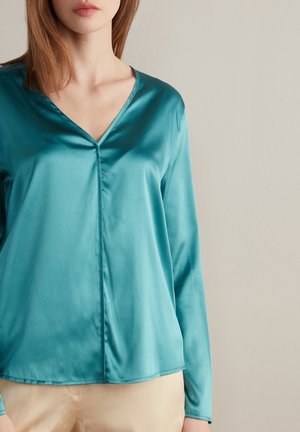 Vrouw in een teal satin blouse met V-hals en lange mouwen, met beige broek, staand tegen een effen lichtgrijze achtergrond.