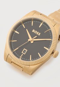 Ρολόι χειρός σε χρυσή απόχρωση με μαύρο καντράν, χρυσά σημάδια ωρών, ένδειξη ημερομηνίας στις 6 η ώρα και το λογότυπο "BOSS" κάτω από τις 12 η ώρα.