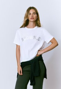 Camiseta blanca de manga corta con el texto plateado brillante "BROWNIE" en el pecho. Tejido texturizado, corte relajado, combinada con pantalones verdes y un suéter oscuro.