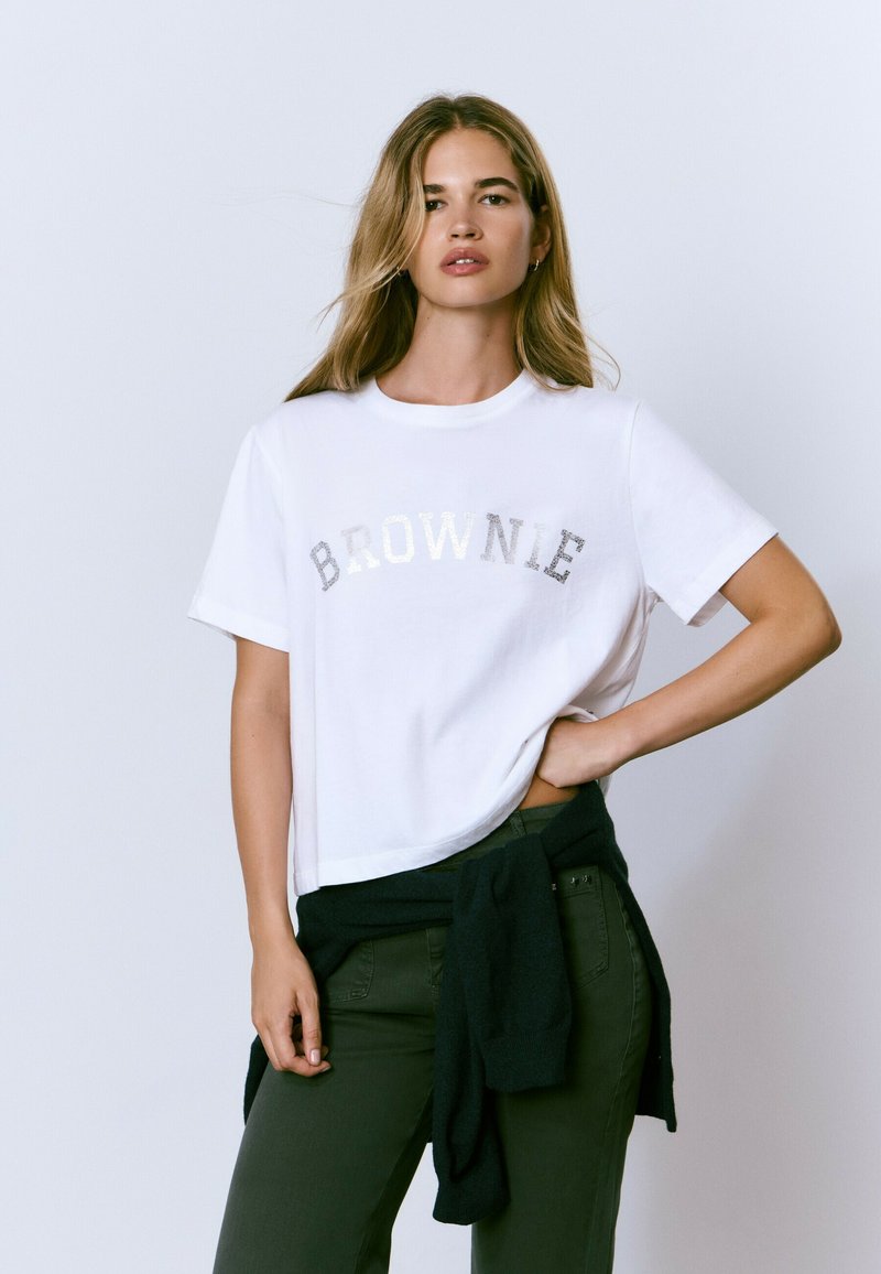 Camiseta blanca de manga corta con el texto plateado brillante "BROWNIE" en el pecho. Tejido texturizado, corte relajado, combinada con pantalones verdes y un suéter oscuro.