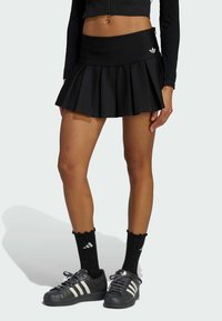 Čierna plisovaná sukňa s priliehavým pásom; skombinovaná s čiernymi športovými topánkami s bielymi pruhmi a čiernymi ponožkami s vlnitým okrajom.