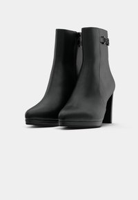 Bottes en cuir noir jusqu'à la cheville avec hauts talons carrés, zips latéraux et boucles décoratives sur un fond blanc.