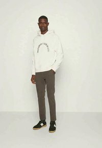 Sweatshirt off-white com um logótipo preto curvado, calças cinzentas justas e sapatilhas pretas com pormenores brancos e solas de borracha. Design simples, estilo casual.