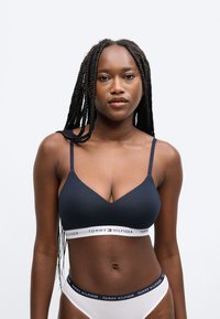 Námornícky modrý bralette s tenkými ramienkami a bielym pásom so značkou. Vysoký pás bielej spodnej bielizne s námornícky modrým pásom. Hladká textúra, minimalistický dizajn.