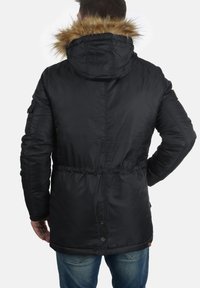Veste matelassée noire avec une capuche doublée en fourrure, taille cintrée, poches latérales et fermeture à boutons-pression. Tissu texturé avec une finition lisse.