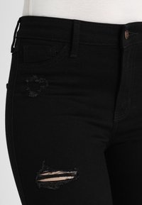 Černé denimové džíny se slim fit střihem, s odřenými záplatami na stehnech a tradičním pětikapsovým designem. Zapínání na knoflík v pase.