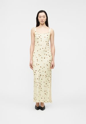 Γυναίκα που στέκεται, φοράει μακρύ κρεμ φόρεμα με καφέ βούλες και μαύρα μυτερά παπούτσια, πάνω σε απλό ανοιχτόχρωμο φόντο.