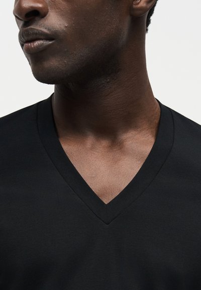 Κοντινό σε κάτω μέρος προσώπου και λαιμό άνδρα που φοράει μαύρο μπλουζάκι με V-λαιμόκοψη σε φωτεινό φόντο.